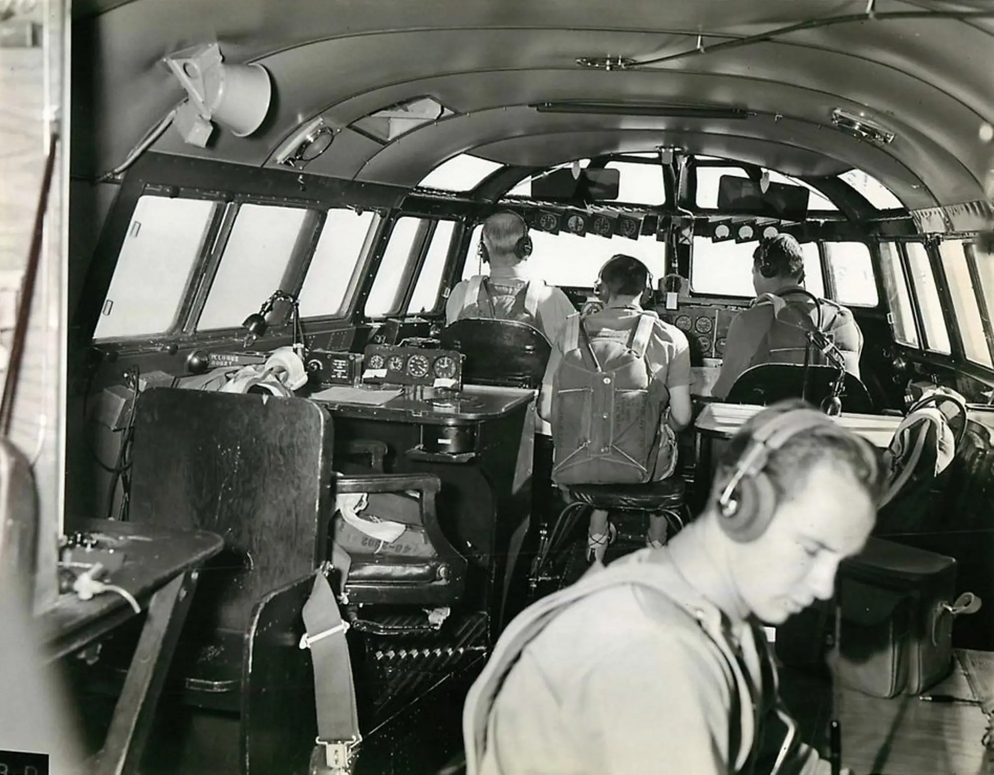 XB-19 “captain’s bridge” during the first flight. Left foreground: empty flight-engineer station with loudspeaker above. The engineer rides a folding seat between the pilots. Major Stanley Umstead occupies the left pilot seat; behind him are the navigator’s table and instruments. Behind the co-pilot is the aircraft commander’s station—here doubling as photographer. Right foreground: radio operator.